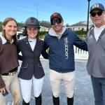 Two women and two men stand on a mat, in front of plastic barricades with structures in the background.