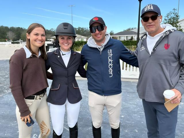 Two women and two men stand on a mat, in front of plastic barricades with structures in the background.