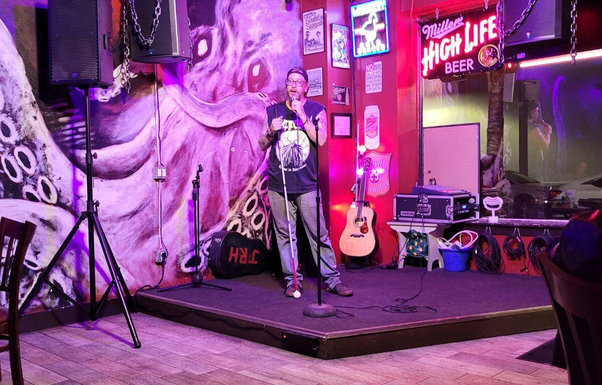 A comedian wearing a cap and light shirt performs on stage, microphone raised, with the octopus mural and neon signage behind them.