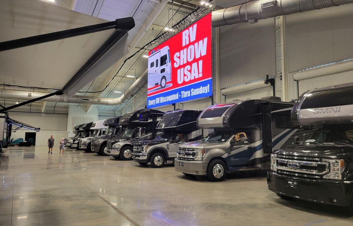 Rows of motorhomes lined up indoors under bright lights at an RV show, with a large “RV Show USA” sign hanging overhead.