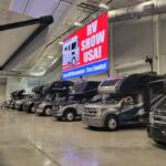 Rows of motorhomes lined up indoors under bright lights at an RV show, with a large “RV Show USA” sign hanging overhead.