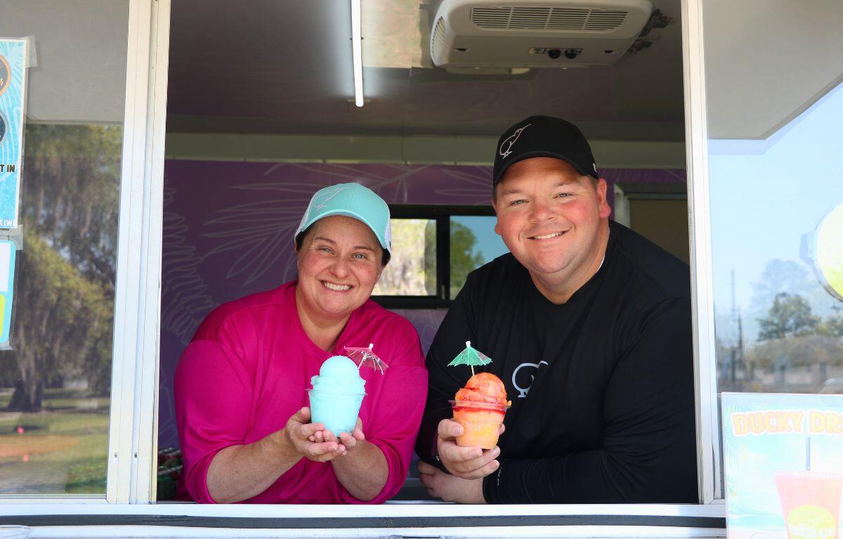 Two people stand in the window of a food truck, holding blue and orange Italian Ice.