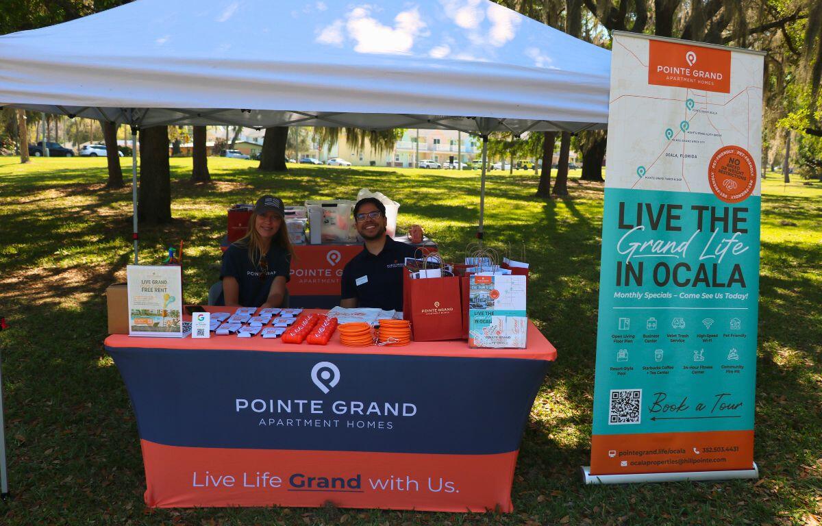 Two people sit together at a table under a white canopy. The tablecloth is orange and gray reading, "Pointe Grand." On the table is advertorial items.