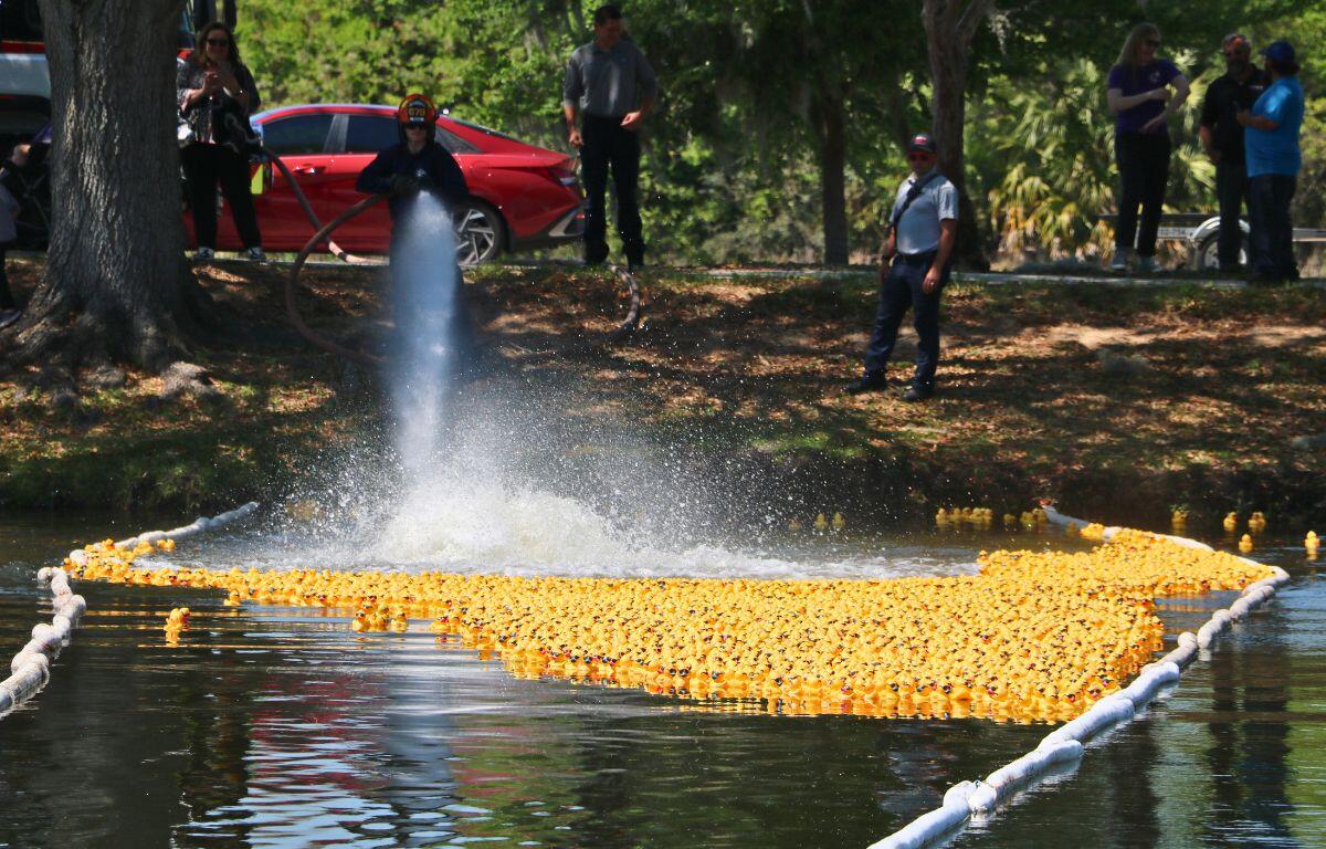 Hundreds of rubber ducks float along a river path marked by a white floating device. A firefighter sprays a strong hose to move them.