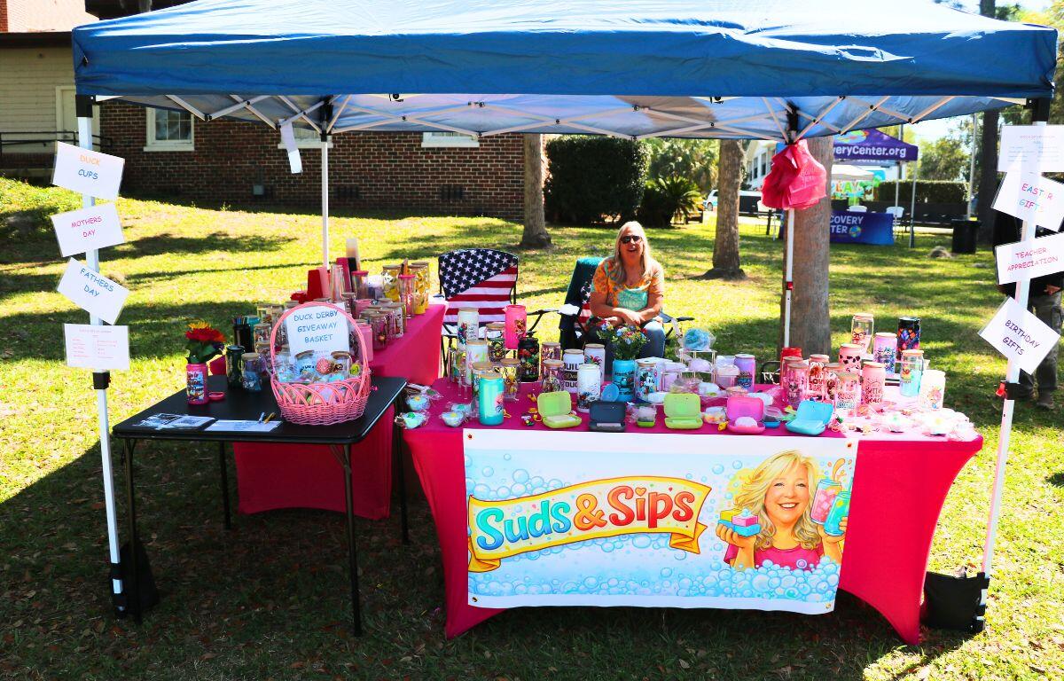 A woman sits behind a table under a canopy with multiple items for sale and giveaways.