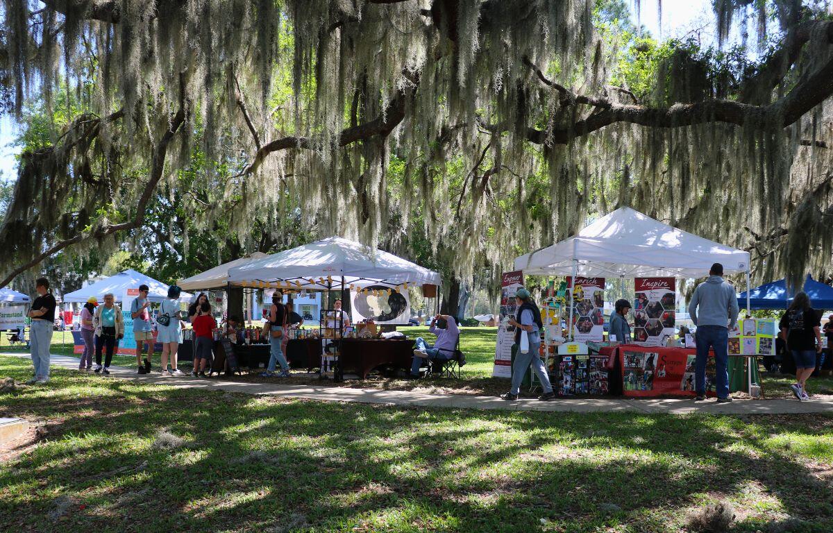 Canopies are lined along a sidewalk shaded by tall trees. Under the canopies are vendors selling items.
