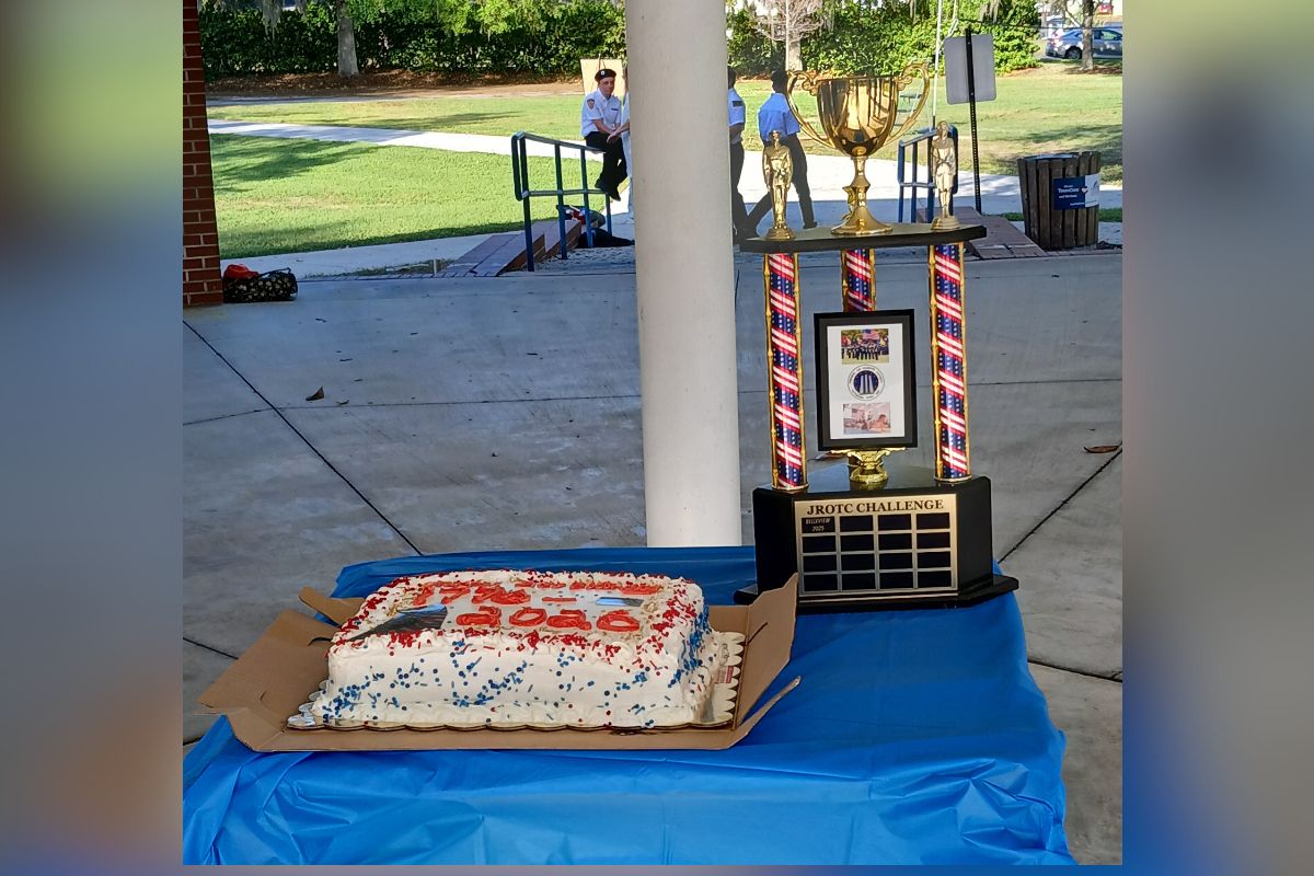 A blue tarp on a table in the foreground, with a cake and trophy, with a column in the foreground, pavement, green space, and people in the background. 