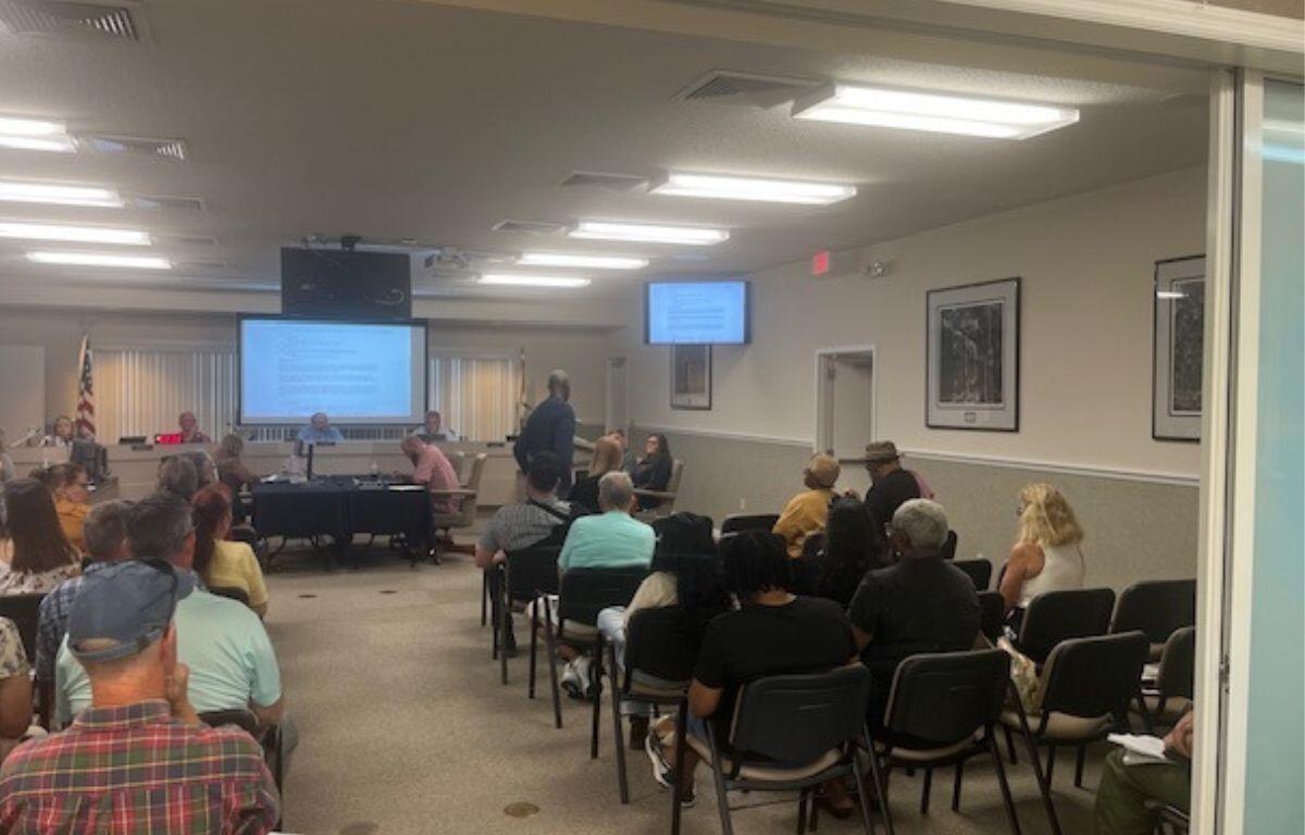 In the foreground people sitting in chairs, while moving toward the background, there are tables with multiple people sitting in front of them, and behind them there is a dais with people sitting behind that and a huge monitor, behind the people. Toward the right there's another table toward the wall, and in front of that a man is in front of a podium with a microphone.