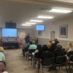 In the foreground people sitting in chairs, while moving toward the background, there are tables with multiple people sitting in front of them, and behind them there is a dais with people sitting behind that and a huge monitor, behind the people. Toward the right there's another table toward the wall, and in front of that a man is in front of a podium with a microphone.