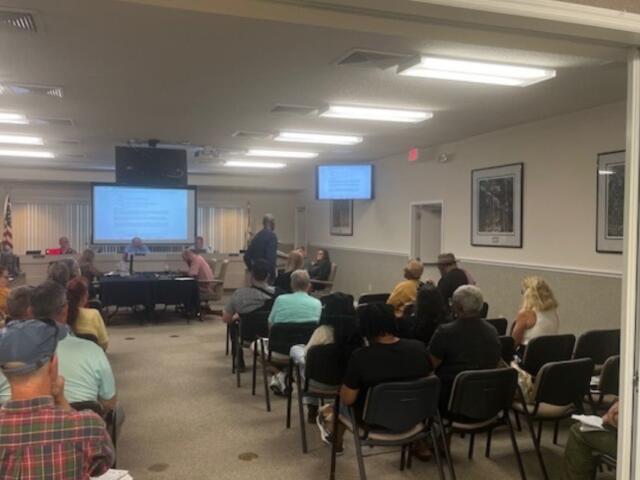 In the foreground people sitting in chairs, while moving toward the background, there are tables with multiple people sitting in front of them, and behind them there is a dais with people sitting behind that and a huge monitor, behind the people. Toward the right there's another table toward the wall, and in front of that a man is in front of a podium with a microphone.