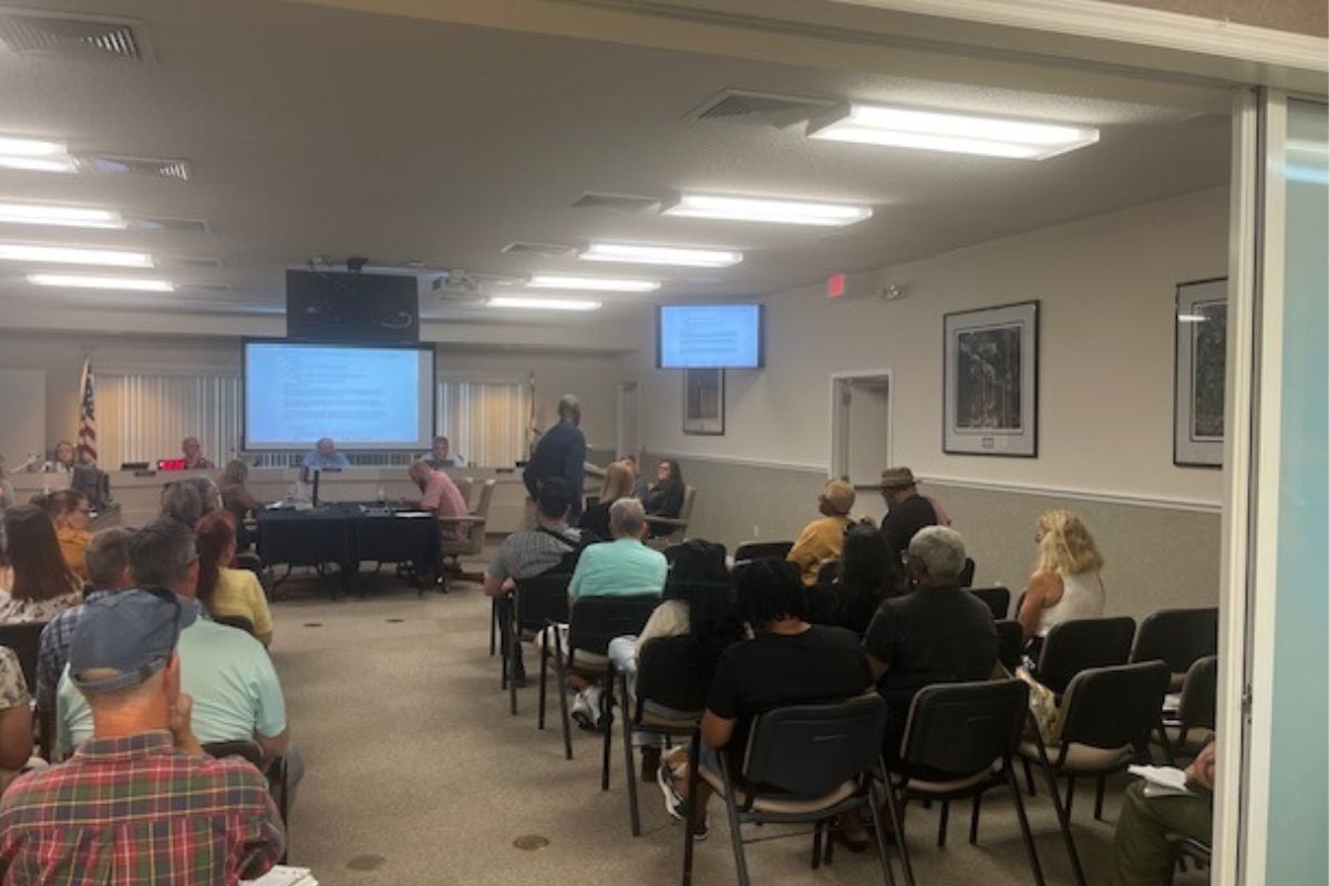 In the foreground people sitting in chairs, while moving toward the background, there are tables with multiple people sitting in front of them, and behind them there is a dais with people sitting behind that and a huge monitor, behind the people. Toward the right there's another table toward the wall, and in front of that a man is in front of a podium with a microphone.