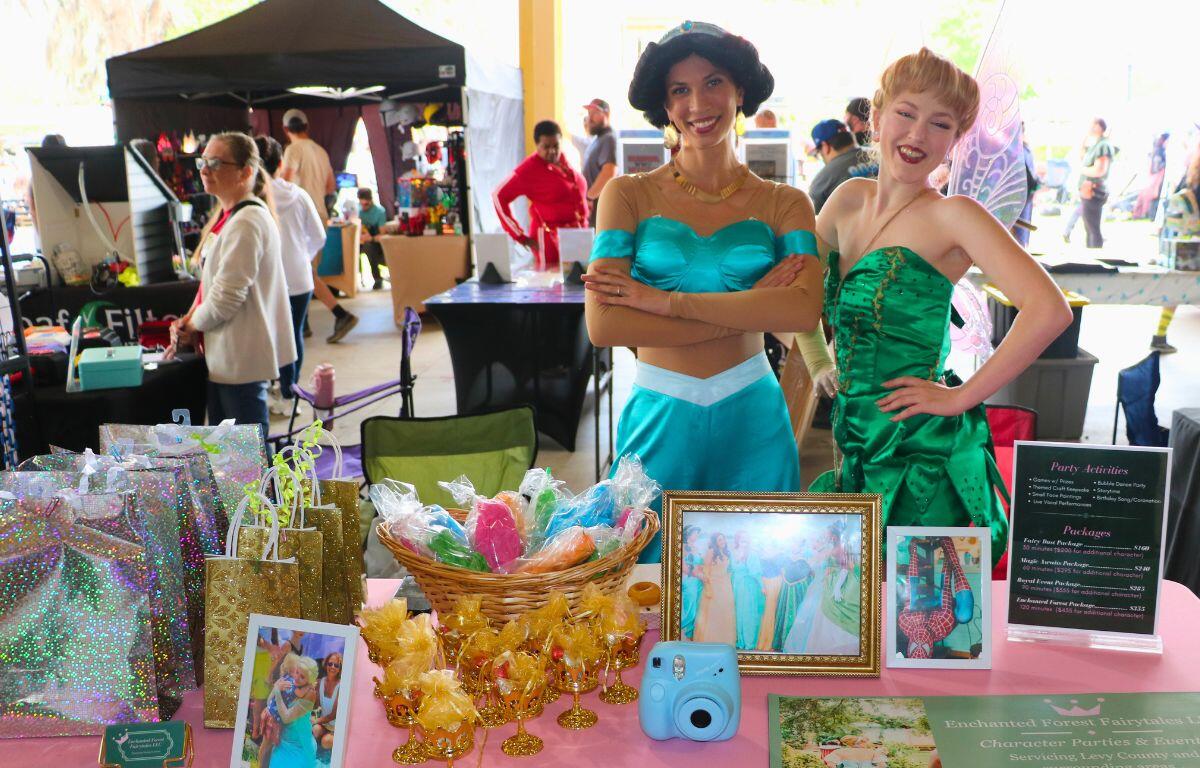 Two women stand behind a table with fantasy items from fairytales. One woman is dressed as Jasmin from "Aladdin."