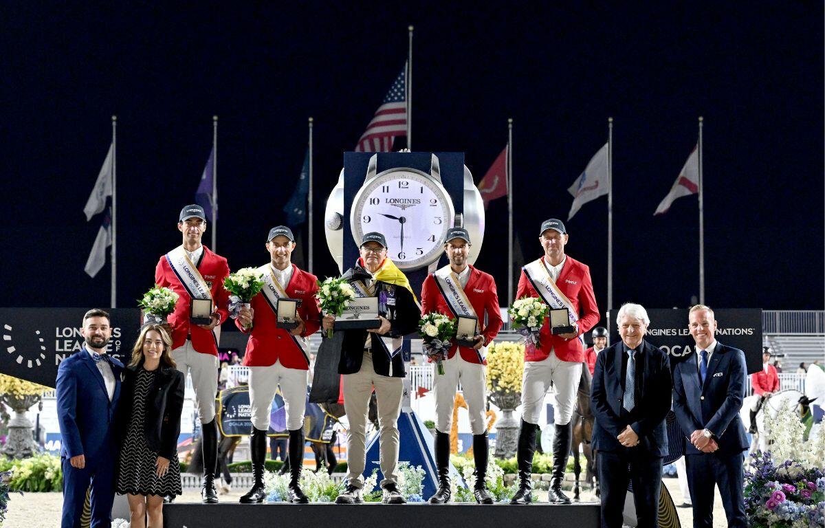 A dark sky, large clock and flags in the background. Men in red blazers and sashes standing on the podium in the middle and people on the left and right of the podium in the foreground.