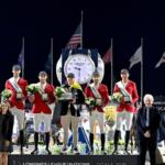 A dark sky, large clock and flags in the background. Men in red blazers and sashes standing on the podium in the middle and people on the left and right of the podium in the foreground.