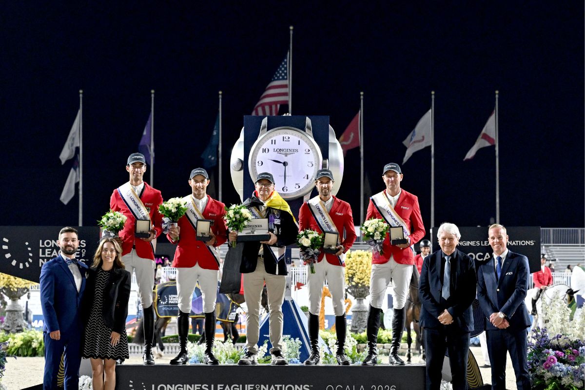 A dark sky, large clock and flags in the background. Men in red blazers and sashes standing on the podium in the middle and people on the left and right of the podium in the foreground.