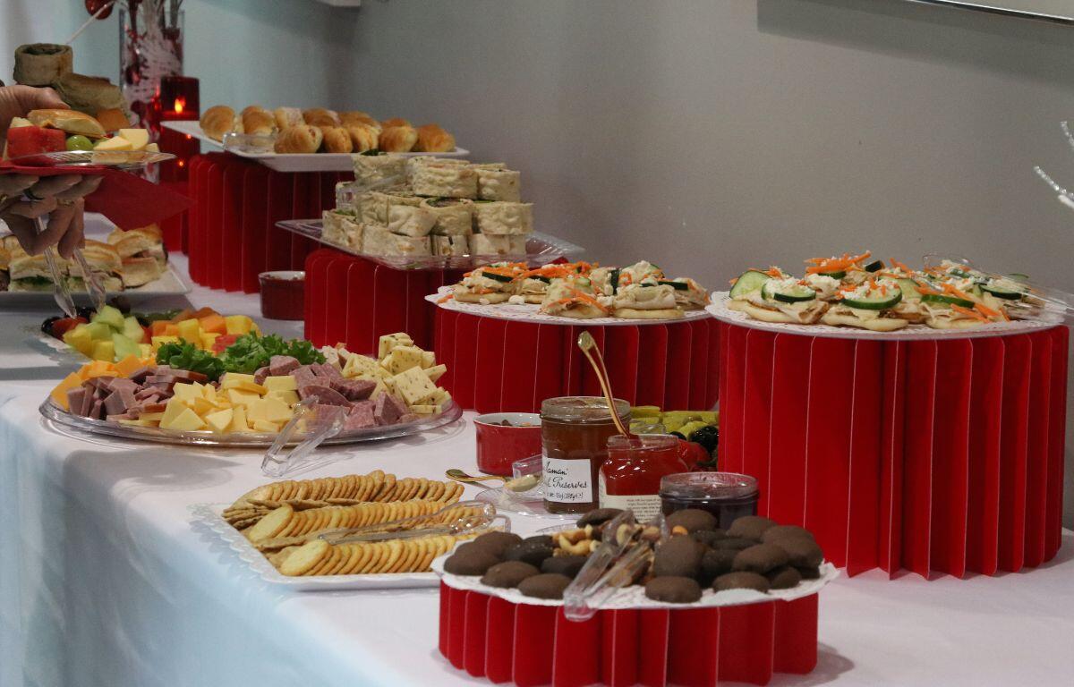 A long table features charcuterie items including crackers, cheese, meat cubes, fruits, small deserts and more. Some items are on higher tiers on top of red table toppers.