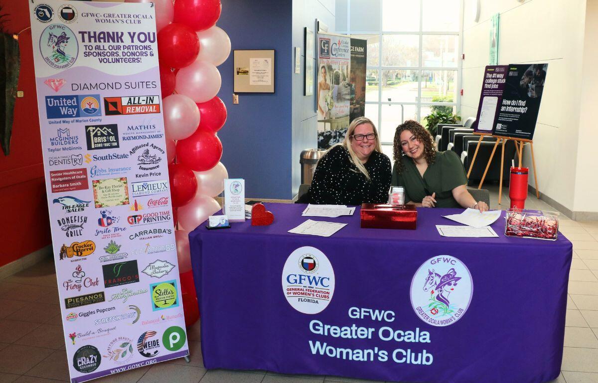 A table with a pink tablecloth features two logos on the front advertising the Greater Ocala Woman's Club with a standing banner to the left reading out a thank you to donors for an event.