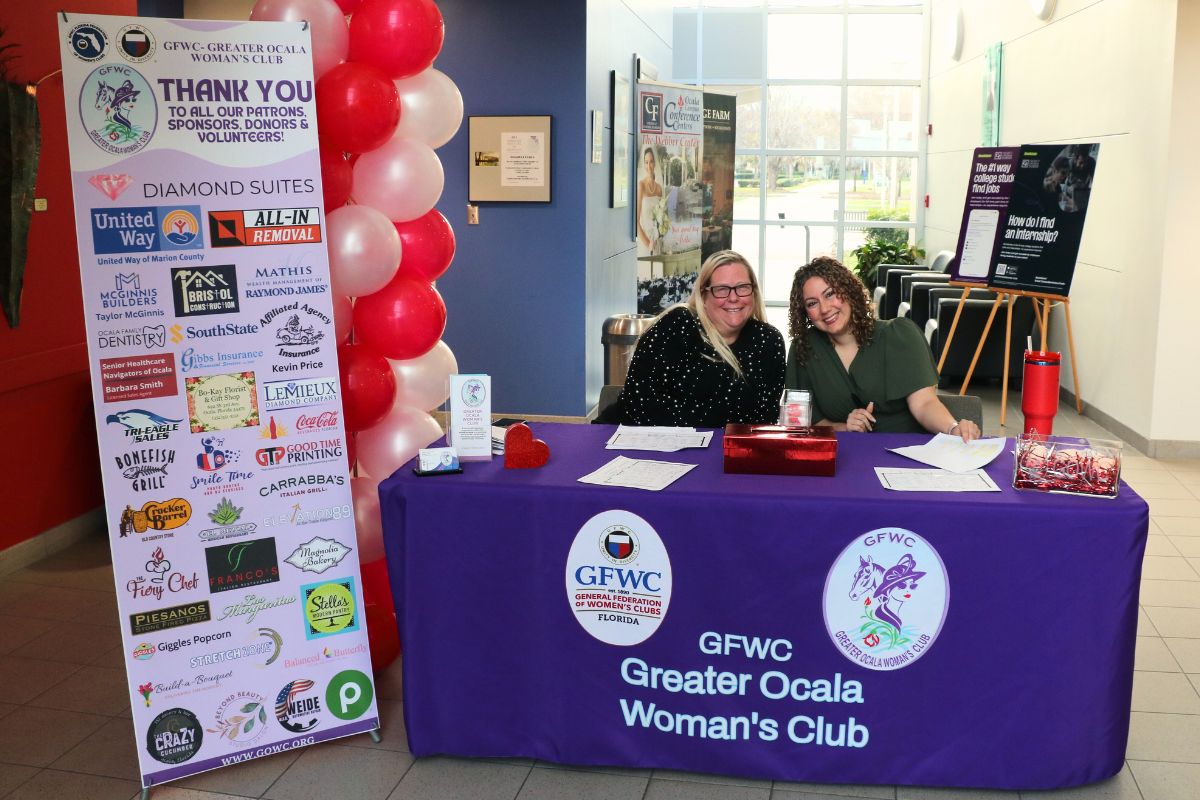 A table with a pink tablecloth features two logos on the front advertising the Greater Ocala Woman's Club with a standing banner to the left reading out a thank you to donors for an event.