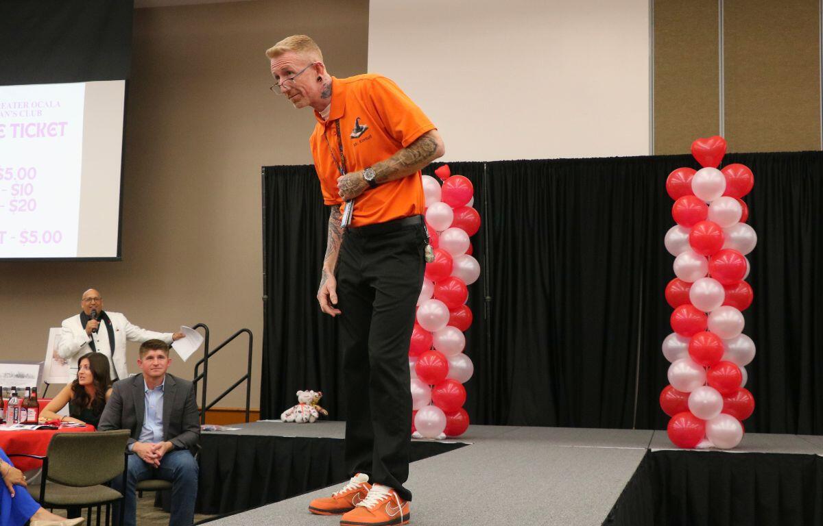A man stands at the end of a catwalk, leaning forward slightly with his glasses slightly adjusted downward while holding onto a lanyard. Surrounding him is a crowd of people watching and a man in the background announcing him.