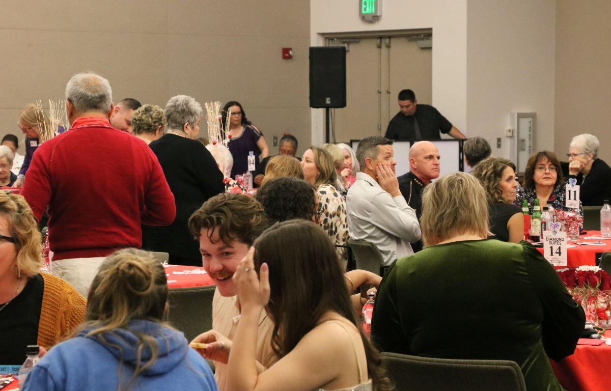 A crowd of people sit at tables and stand in circles, conversing during an event. A DJ stands in the background with a speaker to his right.
