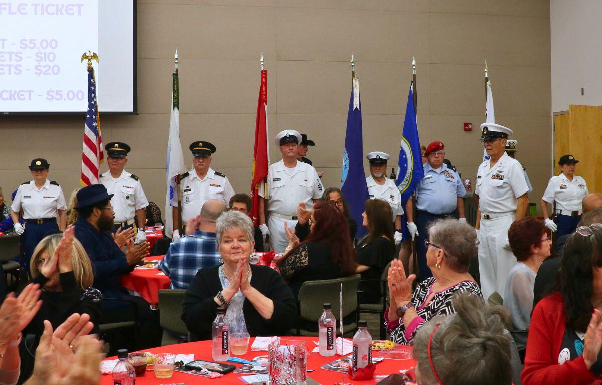 A line of people dressed in honor guard uniforms and holding flags stand facing a crowd. The crowd of guests clap as the honor guard took their positions.