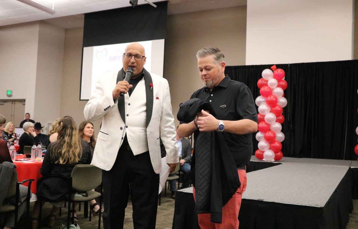 Two men stand together in front of a catwalk. One man is speaking into a microphone while the other holds a jacket. In the background is a group of guests watching.