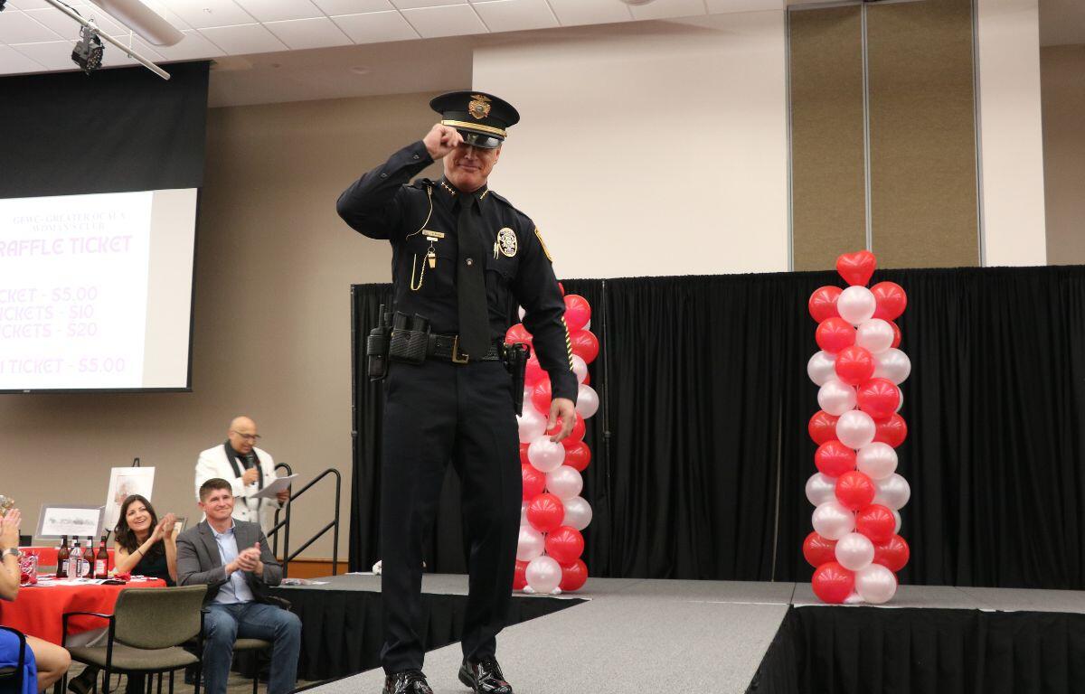 A man in a police chief uniform stands at the end of a catwalk, saluting his hat to a crowd of people applauding.