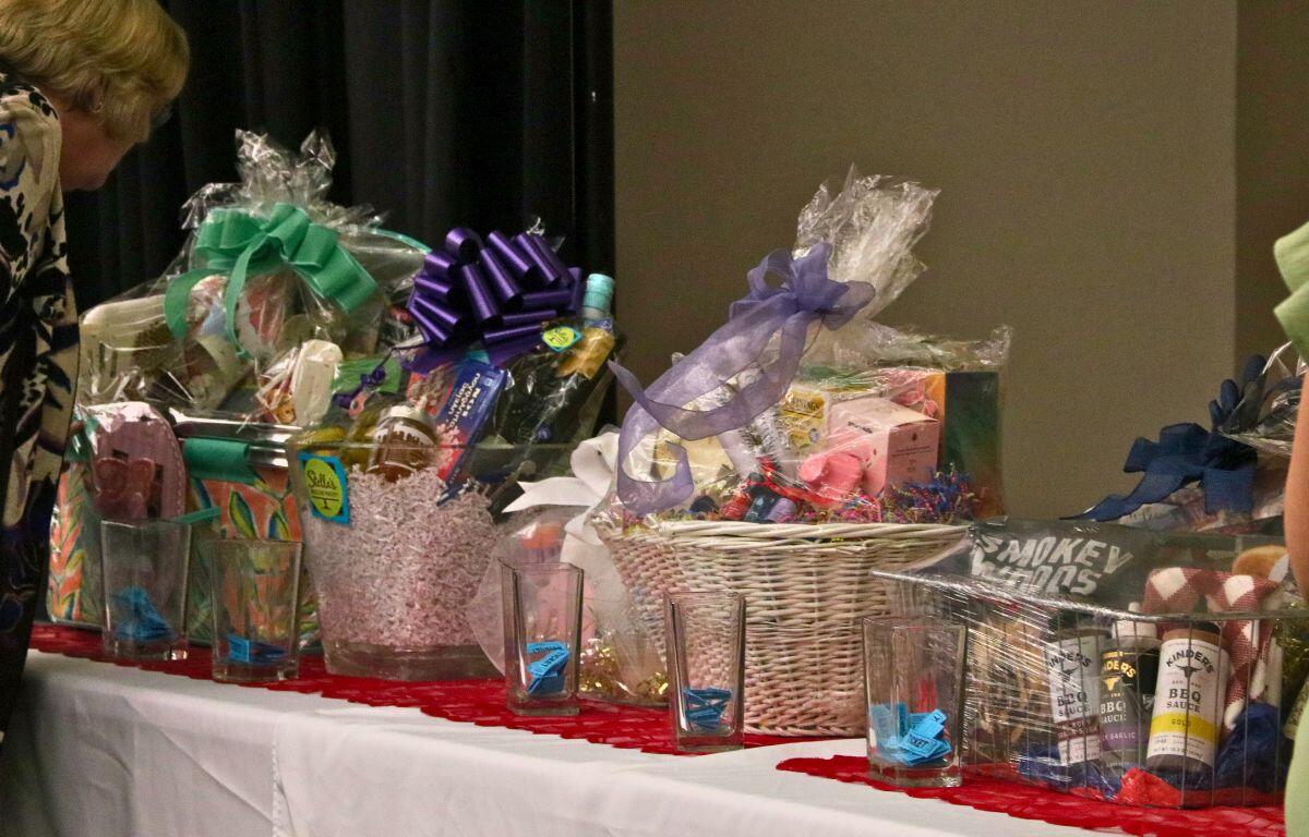 A table is lined with gift baskets with small cups of raffle tickets in front of them. Guests peer at the baskets, placing raffle tickets into the cups.