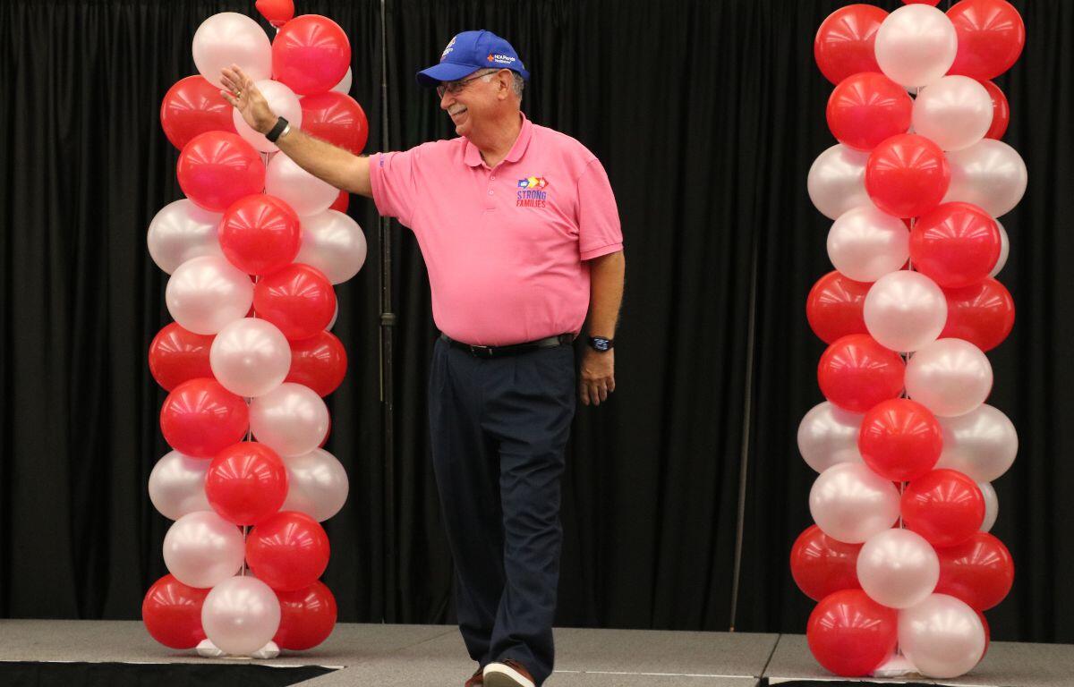 A man walks down a catwalk, waving to a crowd of people with decorative red and white balloons on either side of him.