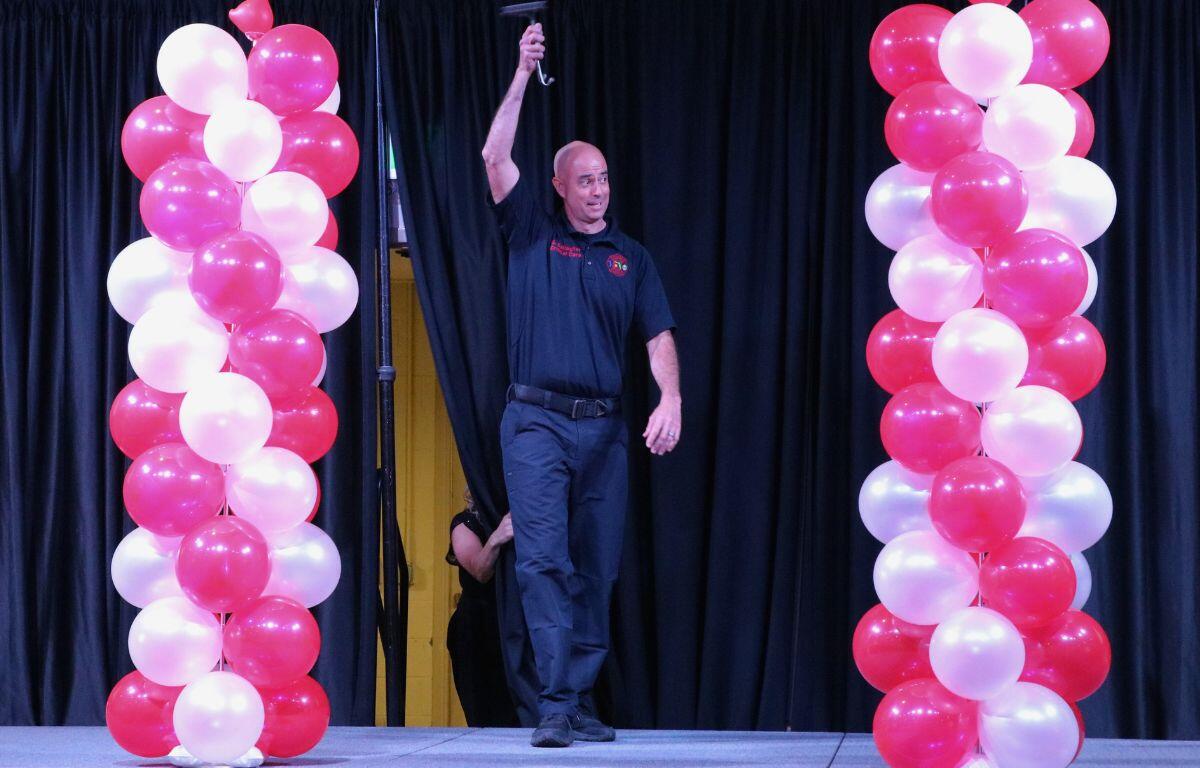 A man in a critical care, emergency service shirt waves a stethoscope in the air while walking down a catwalk with red and white balloons on either side of him.