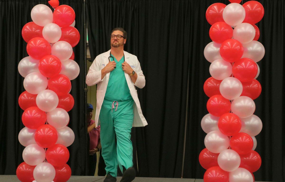A man wearing green scrubs, a doctor coat and a stethoscope walks down a catwalk with red and white balloons on either side of him.