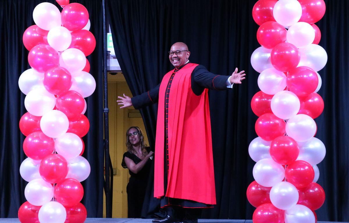 A man in a priests outfit poses on a stage with red balloons on either side of him and a black curtain being held slightly open behind him.