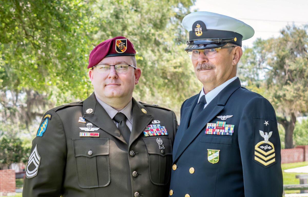 On the left a man dressed in a U.S. Army uniform wearing a beret and to his left a man dressed in a U.S. Navy uniform with trees in the background.