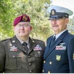 On the left a man dressed in a U.S. Army uniform wearing a beret and to his left a man dressed in a U.S. Navy uniform with trees in the background.