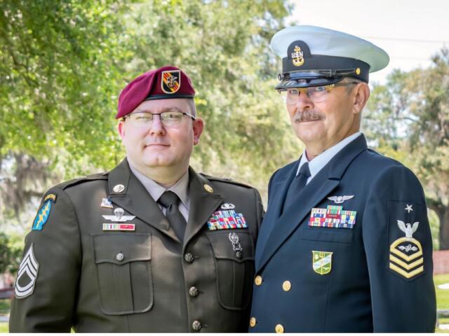 On the left a man dressed in a U.S. Army uniform wearing a beret and to his left a man dressed in a U.S. Navy uniform with trees in the background.