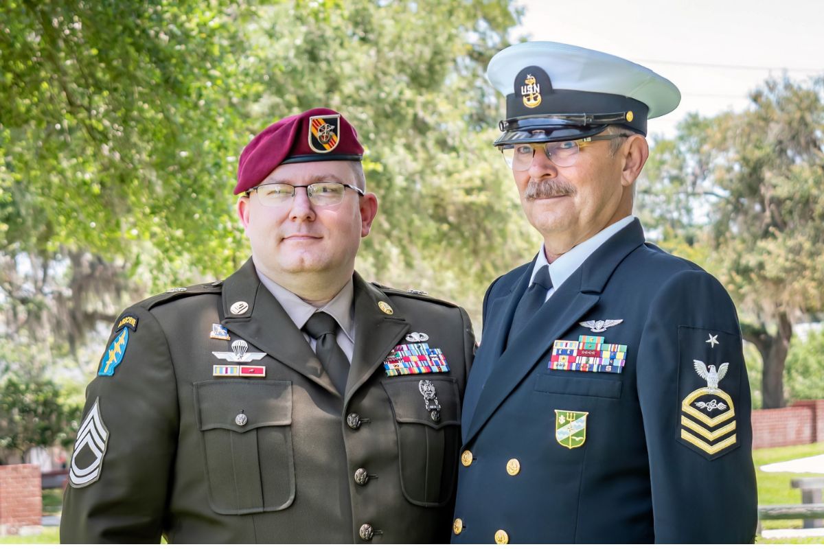 On the left a man dressed in a U.S. Army uniform wearing a beret and to his left a man dressed in a U.S. Navy uniform with trees in the background.