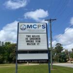 Grass in the foreground, a large sign with a logo and writing, to its immediate right a telephone poll, trees in the background, a hanging street sigh, and cars and building to the right in the background.
