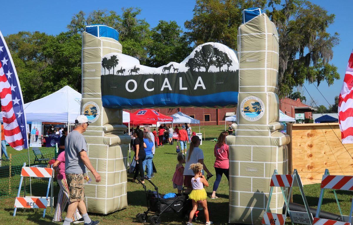 A blow-up arch with the City of Ocala logo on either side and a display of horses walking through a field toward the top. Guests walk underneath it, walking toward vendors under canopies.