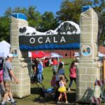 A blow-up arch with the City of Ocala logo on either side and a display of horses walking through a field toward the top. Guests walk underneath it, walking toward vendors under canopies.