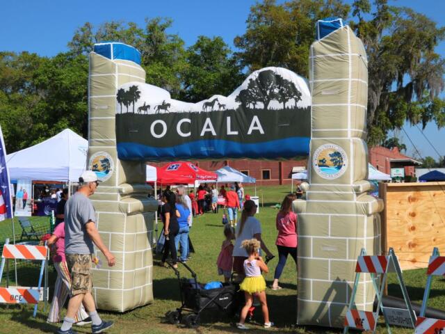A blow-up arch with the City of Ocala logo on either side and a display of horses walking through a field toward the top. Guests walk underneath it, walking toward vendors under canopies.