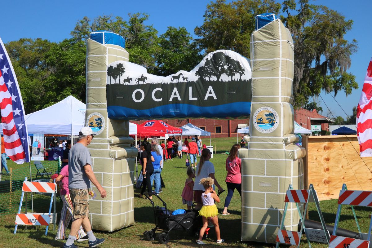 A blow-up arch with the City of Ocala logo on either side and a display of horses walking through a field toward the top. Guests walk underneath it, walking toward vendors under canopies.