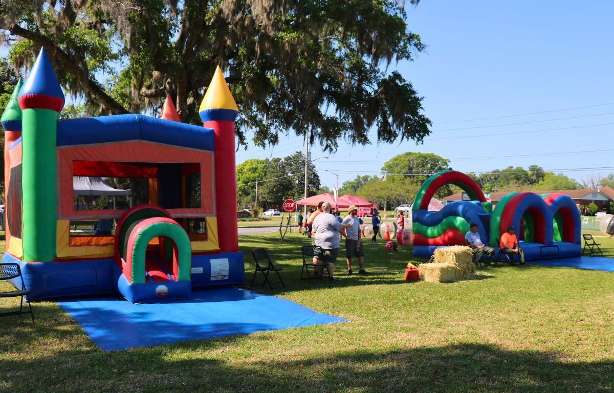 Bounce houses are in a grass field with blue tarps in front of them. People sit around the inflatables while kids play inside.