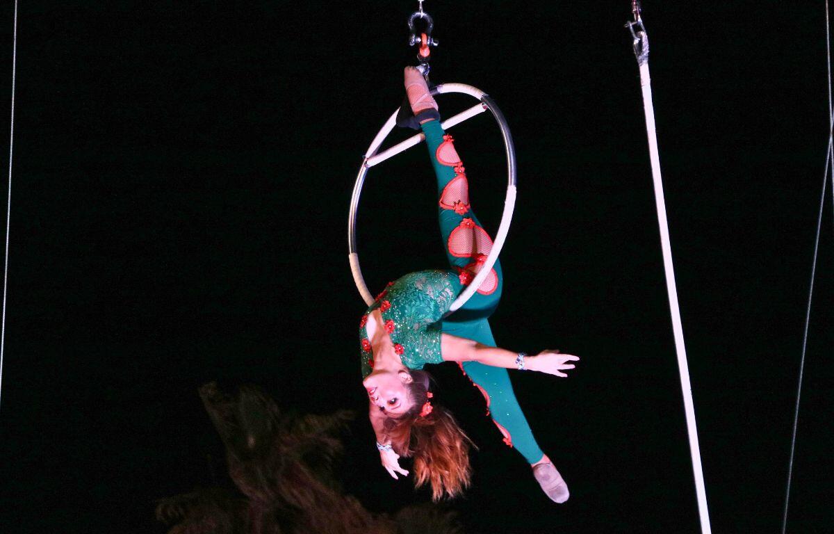 A woman hangs upside down in a vertical split on a hoop lifted in the air.