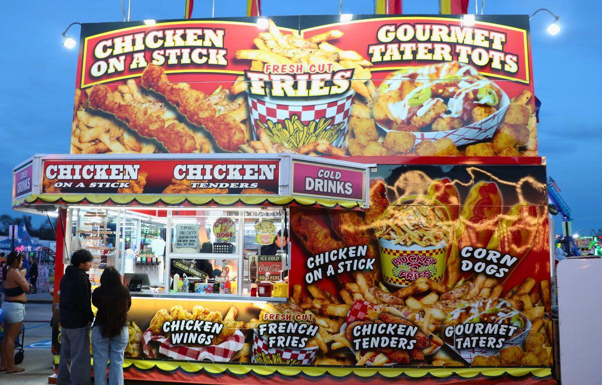A large food vendor features depictions of fries and chicken on its wall. Guests stand at an order window, waiting for their food.