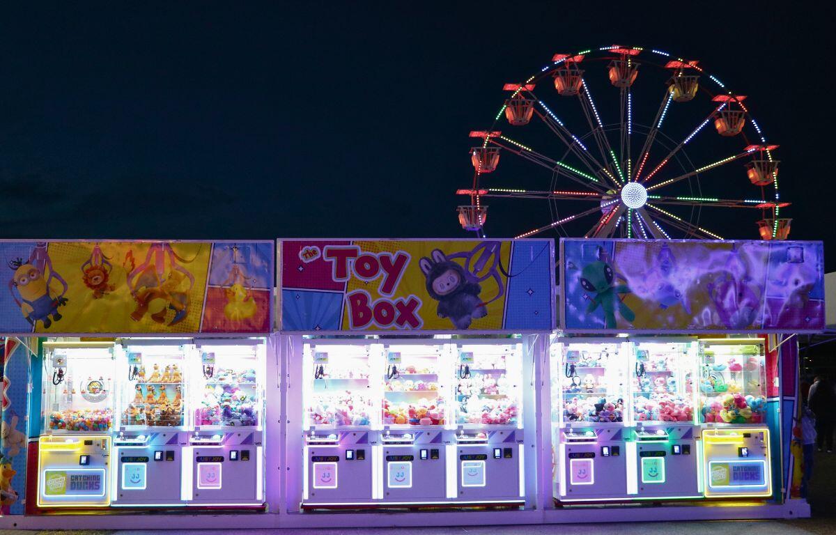 Nine claw machines are lined up together with pictures of prizes at the top of the machines. A large Ferris Wheel with red, green, blue and yellow lights stand in the background.