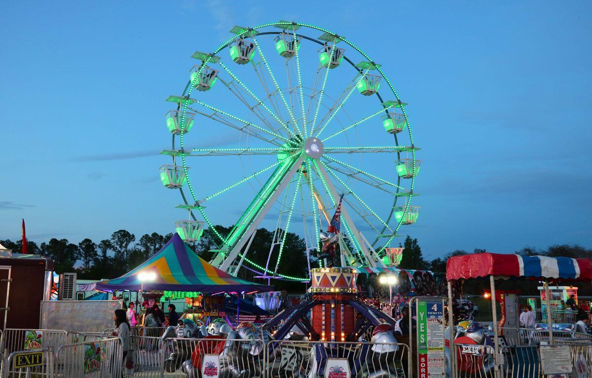 A large Ferris Wheel stands behind kids' rides at a fair as the sun sets.