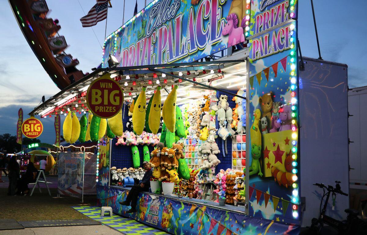 A balloon popping fair game features a large booth with a name at the top reading, "Chloe's Prize Palace." Stuffed animals are hung around the booth.