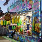 A balloon popping fair game features a large booth with a name at the top reading, "Chloe's Prize Palace." Stuffed animals are hung around the booth.