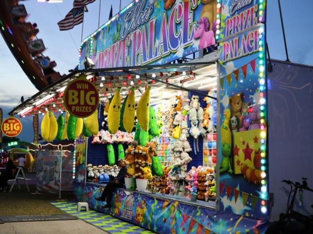 A balloon popping fair game features a large booth with a name at the top reading, "Chloe's Prize Palace." Stuffed animals are hung around the booth.