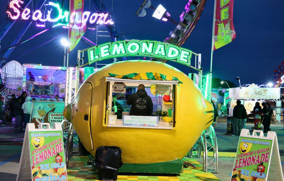 A lemonade stand is bright yellow and in the shape of a lemon. Above it is a neon sign that reads, "Lemonade." In the background is a lively fair.